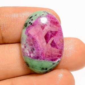 May include: An oval gemstone displaying a striking contrast of colours. The central area is a vivid pink, with a crystalline pattern, encircled by a green band speckled with black. The stone is held in a hand.