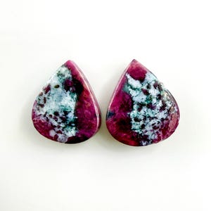 May include: Two teardrop-shaped gemstones with a vibrant color palette. The stones feature a deep magenta base, contrasted by patches of white and teal, creating a marbled effect. The polished surfaces reflect light, highlighting the natural patterns.