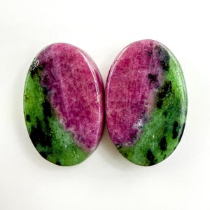 May include: Two oval gemstones. Each stone displays a striking colour combination: a central area of deep pink or magenta, encircled by a band of green with black inclusions. The stones are polished and smooth.