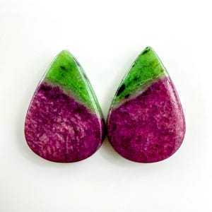 Puede incluir: Dos piedras preciosas en forma de lágrima. La parte superior de cada piedra es de un verde vibrante, que pasa a un tono magenta intenso en la mitad inferior. Las piedras tienen una superficie pulida y lisa, sobre un fondo blanco.