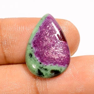 May include: A teardrop-shaped gemstone with a polished surface. The stone features a vibrant mix of colors, including a deep magenta center, surrounded by a green border with black speckles. The gemstone is held in a hand.