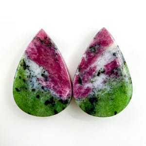 Może przedstawiać: Dwa kamienie szlachetne w kształcie łezki o żywej palecie barw. Kamienie mają gradient od zieleni u podstawy, przechodzący w biel, a następnie w głęboki róż na górze. Czarne plamki dodają szczegółów do wypolerowanej powierzchni.