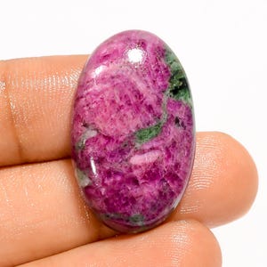 Puede incluir: Una gema de forma ovalada con una superficie pulida. La piedra presenta una vibrante mezcla de tonos rosa y magenta, con vetas verdes y blancas. La gema se sostiene en una mano, mostrando su tamaño y forma.