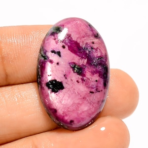 Puede incluir: Una gema ovalada con superficie pulida. La piedra presenta una mezcla de tonos rosa y púrpura oscuro, con inclusiones negras. La gema se sostiene en una mano, mostrando su tamaño y variaciones de color. La piedra mide aproximadamente 3 cm de largo.