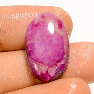 Könnte beinhalten: Ein polierter, oval geformter Edelstein mit einer lebendigen Farbpalette aus Pink und Magenta. Der Stein hat eine transluzente Qualität mit inneren Mustern und Einschlüssen, die eine einzigartige visuelle Textur erzeugen. Der Edelstein wird in einer Hand gehalten.