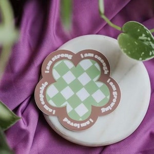 May include: A brown flower-shaped sticker with a green and white checkered pattern in the center. The sticker has the text "I am strong, I am capable, I am loved, I am enough, I am growing" written around the edge.