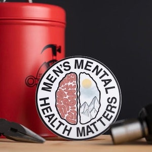 May include: A black and white sticker with the text "Men's Mental Health Matters" in a circle. The sticker features a stylized image of a brain split in half, one side showing a brain and the other side showing a mountain range.