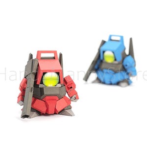 Pu&ograve; includere: Due figurine di robot, una rossa e una blu, sono esposte su sfondo bianco. Il robot rosso &egrave; in primo piano, con una pistola attaccata. Ogni robot ha un globo giallo brillante al centro.