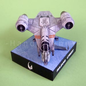 May include: A detailed model of the Razor Crest starship, a spacecraft from a popular science fiction series. The model is primarily gray with orange accents and is mounted on a blue and black base. The base has the text "RAZOR CREST".