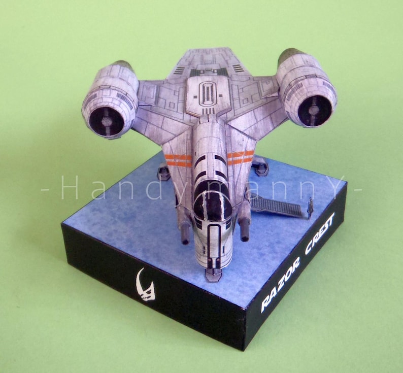 May include: A detailed model of the Razor Crest starship, a spacecraft from a popular science fiction series. The model is primarily gray with orange accents and is mounted on a blue and black base. The base has the text "RAZOR CREST".