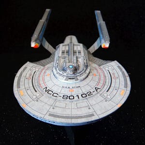 May include: A model of the U.S.S. Titan starship, featuring a gray and white color scheme with red and orange accents. The ship has a saucer-shaped primary hull and two warp nacelles. The text "NCC-80102-A" is visible on the hull.
