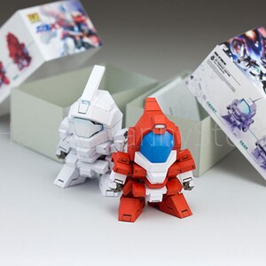 May include: Two small, assembled robot figures, one white and one red with blue accents, stand in front of a gray box. Two boxes with illustrations of robots are behind them. The figures appear to be from a model kit.