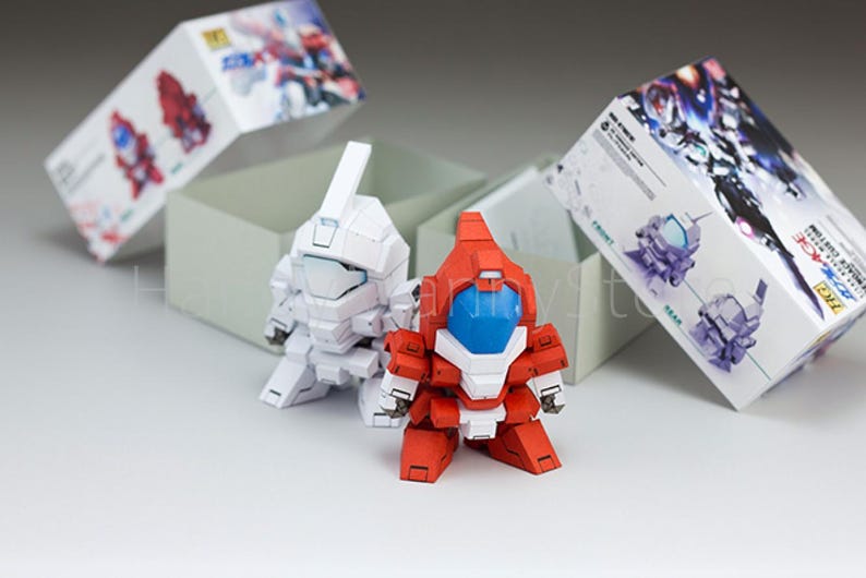 May include: Two small, assembled robot figures, one white and one red with blue accents, stand in front of a gray box. Two boxes with illustrations of robots are behind them. The figures appear to be from a model kit.
