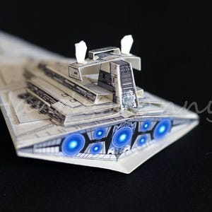 May include: A paper model of a Star Destroyer, with detailed construction and glowing blue engine lights. The model is mainly white with grey detailing, showing a complex, multi-layered structure. The spacecraft is set against a dark background.