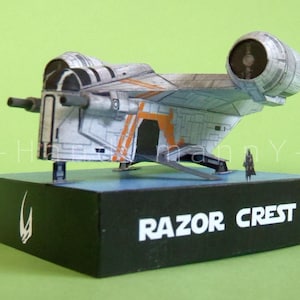 May include: A model of the Razor Crest starship, a detailed replica in shades of gray, white, and orange, mounted on a black base. The base features the ship's name in white text and a symbol. A small figure stands nearby.