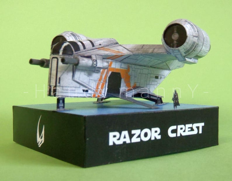 May include: A model of the Razor Crest starship, a detailed replica in shades of gray, white, and orange, mounted on a black base. The base features the ship's name in white text and a symbol. A small figure stands nearby.