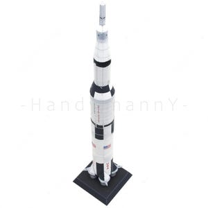 Könnte beinhalten: Ein vertikales Modell der Saturn V-Rakete mit einem weißen zylindrischen Körper, schwarzen Segmenten, einer spitzen Oberseite und einem quadratischen schwarzen Sockel. Die Rakete zeigt den Schriftzug UNITED STATES und USA sowie kleine US-Flaggen.
