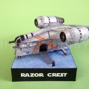 May include: A detailed model of the Razor Crest starship, featuring a gray and silver color scheme with orange accents. The model sits on a blue base with a black box labeled "RAZOR CREST". A small figure stands near the ship.