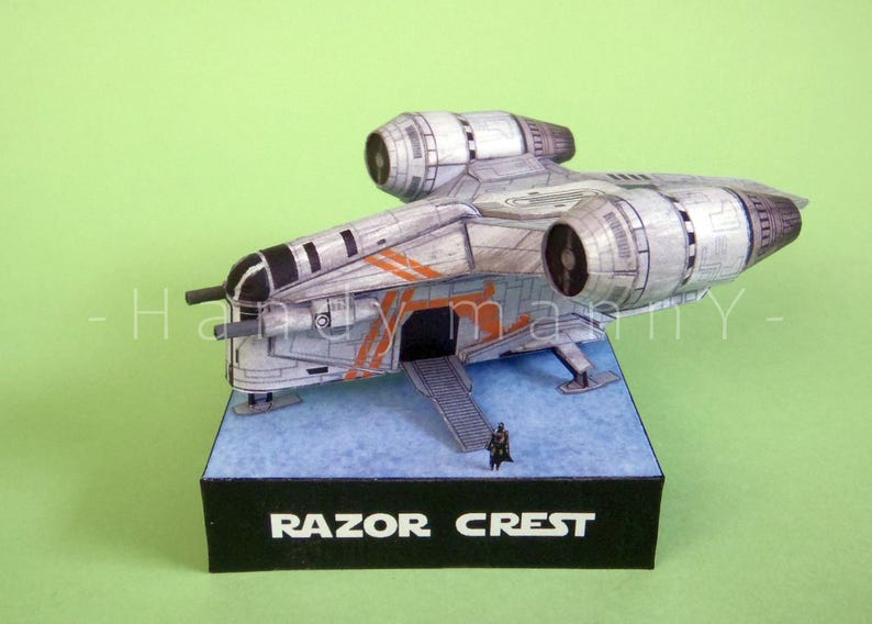 May include: A detailed model of the Razor Crest starship, featuring a gray and silver color scheme with orange accents. The model sits on a blue base with a black box labeled "RAZOR CREST". A small figure stands near the ship.