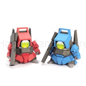Pu&ograve; includere: Due figure simili a robot, una rossa e una blu, sono affiancate su uno sfondo bianco. Ognuna ha un grande occhio giallo brillante e un accessorio a forma di pistola. Le figure sono fatte di carta piegata angolare.