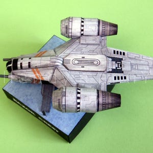 May include: A detailed model of a spacecraft, primarily gray with black and orange accents, is displayed on a blue and black base against a green background. The model features two large engine components and intricate panel detailing.