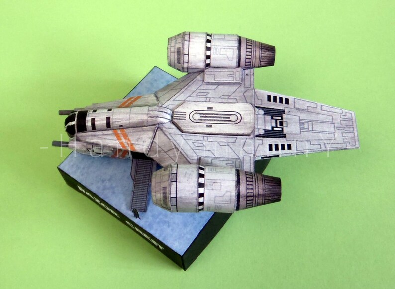 May include: A detailed model of a spacecraft, primarily gray with black and orange accents, is displayed on a blue and black base against a green background. The model features two large engine components and intricate panel detailing.