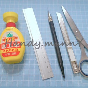May include: A collection of craft tools including tweezers, a yellow bottle with red accents, a metal ruler, a black pen, a utility knife, and scissors. The tools are arranged on a blue grid surface.