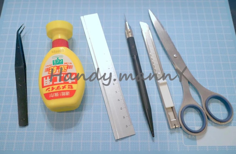 May include: A collection of craft tools including tweezers, a yellow bottle with red accents, a metal ruler, a black pen, a utility knife, and scissors. The tools are arranged on a blue grid surface.