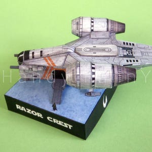 May include: A detailed model of the Razor Crest starship, a spacecraft from a popular science fiction series. The model is primarily gray with orange accents and is mounted on a black base with the words "Razor Crest" printed on it.
