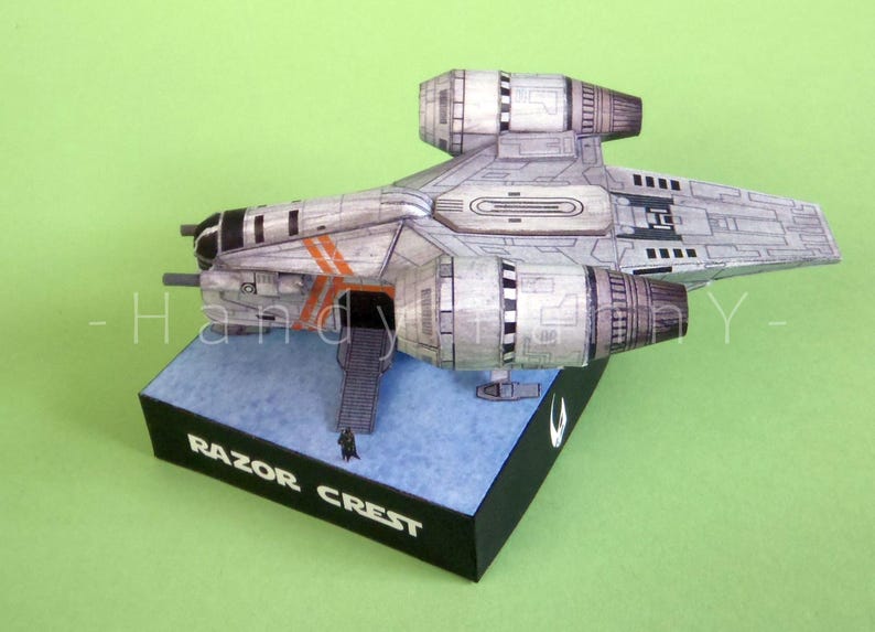 May include: A detailed model of the Razor Crest starship, a spacecraft from a popular science fiction series. The model is primarily gray with orange accents and is mounted on a black base with the words "Razor Crest" printed on it.