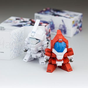 May include: Two small, assembled robot figures, one white and one red, with blue visors, stand on a white surface. Boxes with colorful illustrations and instruction booklets are in the background. The robots have a blocky design.