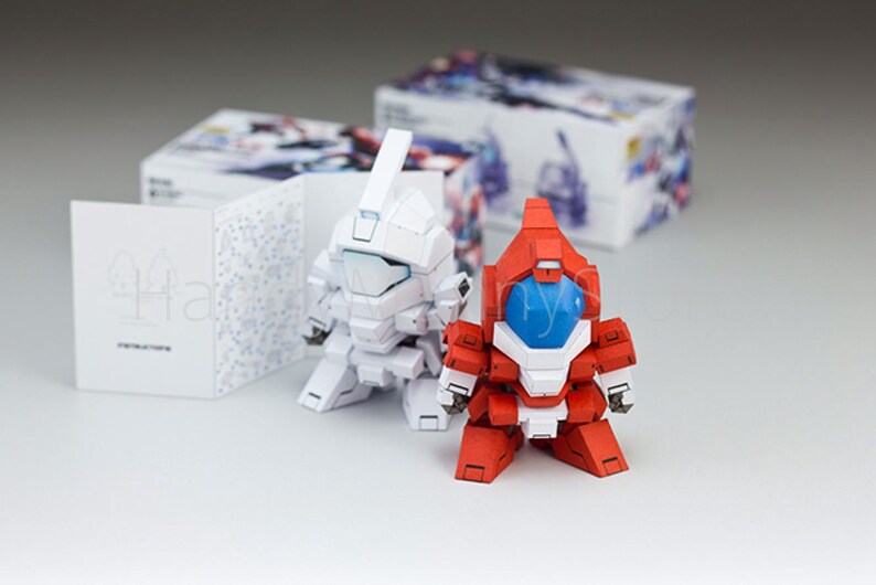 May include: Two small, assembled robot figures, one white and one red, with blue visors, stand on a white surface. Boxes with colorful illustrations and instruction booklets are in the background. The robots have a blocky design.