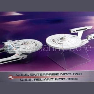 Puede incluir: Dos maquetas de naves espaciales blancas y grises, la U.S.S. Enterprise NCC-1701 y la U.S.S. Reliant NCC-1864, se exhiben en una plataforma morada y gris. La Enterprise está sobre un soporte transparente. La plataforma tiene el texto "U.S.S. ENTERPRISE NCC-1701" y "U.S.S. RELIANT NCC-1864."