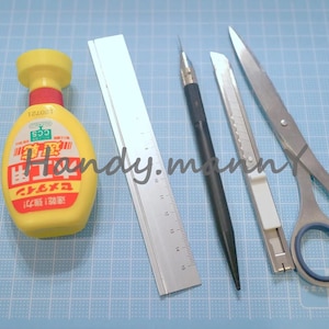 May include: An assortment of craft tools including tweezers, a yellow bottle with Japanese text, a ruler, a black pen, a utility knife, and scissors. The tools are arranged on a blue grid surface.