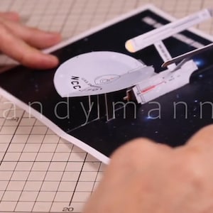 Può includere: Un modello in carta dell'astronave USS Enterprise con una sezione a disco bianca con la scritta NCC-1701, fissato su una base rettangolare nera che raffigura una scena spaziale stellata, assemblato con pinzette su un tappetino a griglia.
