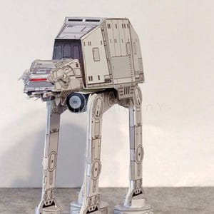 May include: A detailed paper model of an AT-AT walker from Star Wars, with a smaller X-wing fighter model nearby. The AT-AT is primarily white with gray accents, standing on four legs. The X-wing is white and red.
