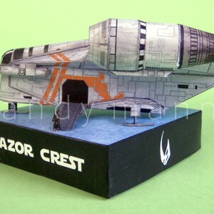 May include: A detailed model of the Razor Crest starship, featuring a gray and orange color scheme. The model sits on a black base with the text "RAZOR CREST" and a white symbol. The background is a solid green.
