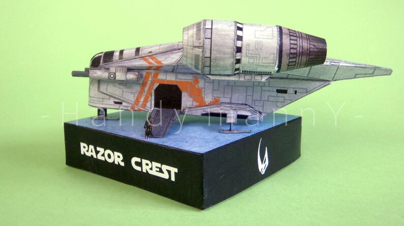 May include: A detailed model of the Razor Crest starship, featuring a gray and orange color scheme. The model sits on a black base with the text "RAZOR CREST" and a white symbol. The background is a solid green.
