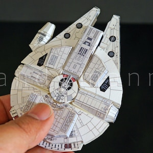 May include: A white and grey paper model of a circular spaceship with intricate paneling details, a central cockpit, and two forward mandibles. The structure features printed mechanical textures and geometric patterns across its flat, layered surface.