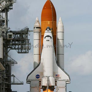 Puede incluir: Una lanzadera espacial blanca con tanques de combustible naranja se lanza desde una plataforma de lanzamiento. La lanzadera está etiquetada con "NASA" y "Atlantis".