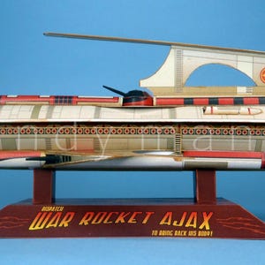 Pode incluir: Um modelo de papel da nave espacial War Rocket Ajax, com um corpo cilíndrico de painéis vermelhos, brancos e bege, uma barbatana superior e uma base de exposição vermelho-escura com o texto "WAR ROCKET AJAX" impresso em letras amarelas.