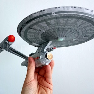 May include: A gray and silver model of the Star Trek USS Enterprise starship. The model has a large saucer section, two warp nacelles, and a secondary hull. The model is held by a hand against a white background.