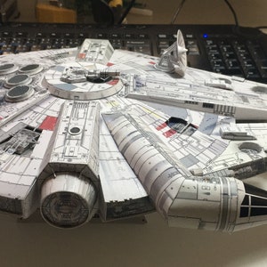 May include: A detailed model of the Millennium Falcon starship, in white and grey with red and black details. The model has intricate paneling and circular details on the top, with a rectangular cockpit and a rear engine.