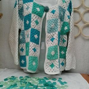 Peut inclure: Un cardigan en crochet blanc avec un motif géométrique bleu et vert. Le cardigan est posé sur une surface blanche avec un tas de carreaux de mosaïque bleus et verts.