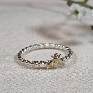 Könnte beinhalten: Ein silberner Ring mit einem gedrehten Band und einem goldenen Bienen-Anhänger.