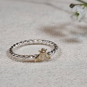 Könnte beinhalten: Ein silberner Ring mit einem gedrehten Band und einem goldenen Bienen-Anhänger.
