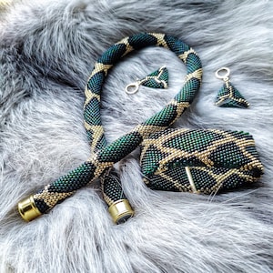 Puede incluir: Un conjunto de joyas de cuentas con collar, pulsera y pendientes. El conjunto es principalmente verde con detalles dorados, creando un patrón geométrico. El collar y la pulsera tienen cierres metálicos dorados. Los pendientes son triangulares.