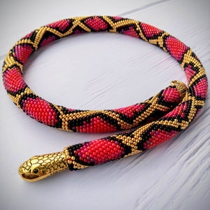 Peut inclure: Un collier de serpent en perles avec un pendentif tête de serpent en or. Le collier est fait de perles rouges, noires et dorées et présente un motif de peau de serpent.