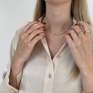 Könnte beinhalten: Eine goldene Kettenkette mit ovalen Gliedern. Die Kette wird von einer Person mit rotem Nagellack und goldenen Ringen an den Fingern getragen.