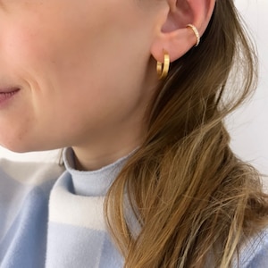 Könnte beinhalten: Eine Frau trägt einen hellblauen Pullover mit einem weißen und hellblauen Muster. Sie trägt zwei goldene Creolen am linken Ohr und einen kleinen goldenen Ohrring mit funkelnden Steinen am rechten Ohr.
